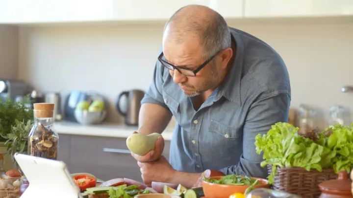 Man holding vegetable, looking at tablet in kitchen